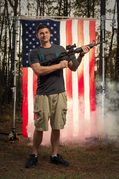 Portrait 3 from the An American Christmas series, person posing with a shotgun in front of an American flag backdrop