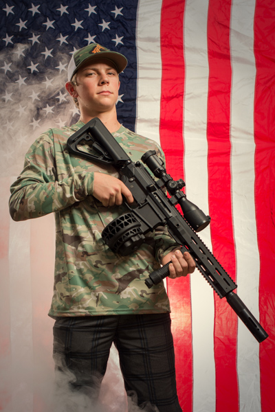 Portrait 7 from the An American Christmas series, person posing with a rifle in front of an American flag backdrop