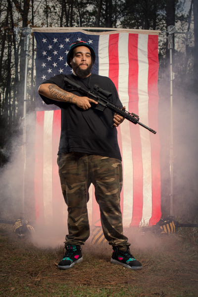 Portrait 13 from the An American Christmas series, person posing with a rifle in front of an American flag backdrop
