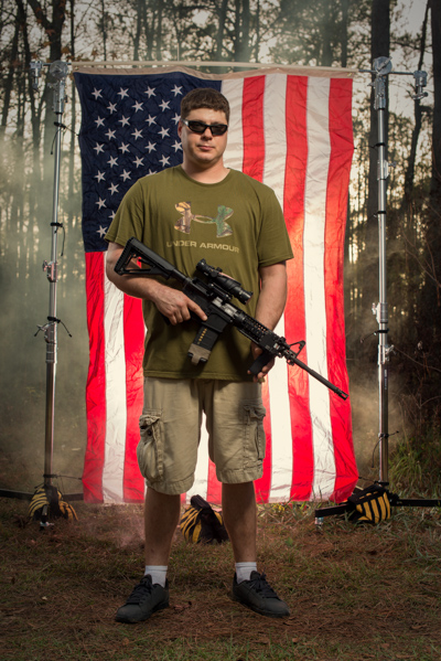 Portrait 14 from the An American Christmas series, person posing with a handgun in front of an American flag backdrop