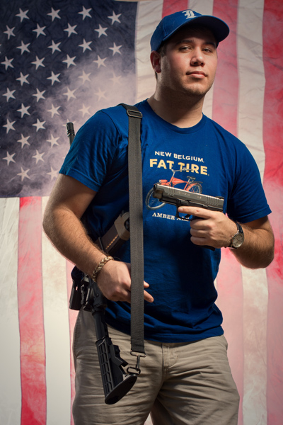 Portrait 21 from the An American Christmas series, person posing with a shotgun in front of an American flag backdrop