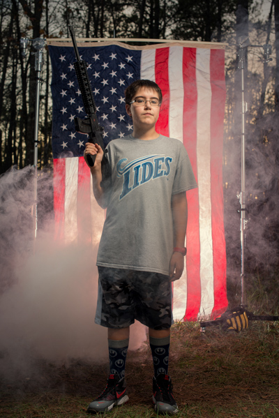 Portrait 23 from the An American Christmas series, person posing with an AR-style rifle in front of an American flag backdrop