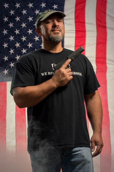 Portrait 25 from the An American Christmas series, person posing with a rifle in front of an American flag backdrop