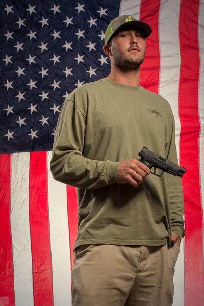 Portrait 28 from the An American Christmas series, person posing with a hunting rifle in front of an American flag backdrop