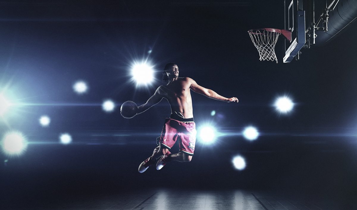 Basketball player dunking, illuminated by camera flashes against a dark gym background