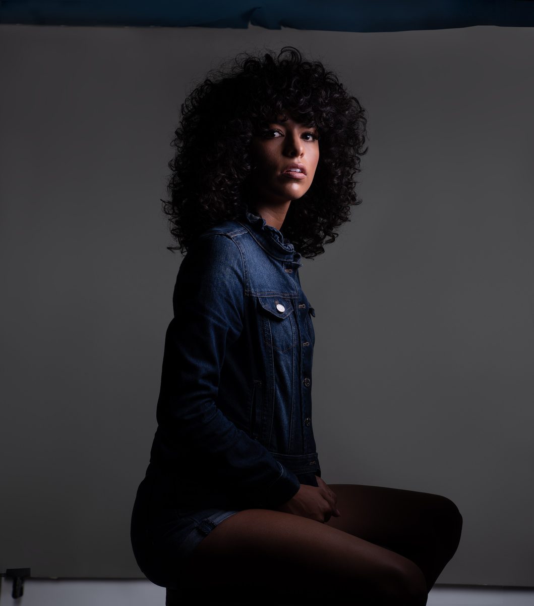 Woman with curly hair wearing a denim jacket, sitting on a stool in a studio