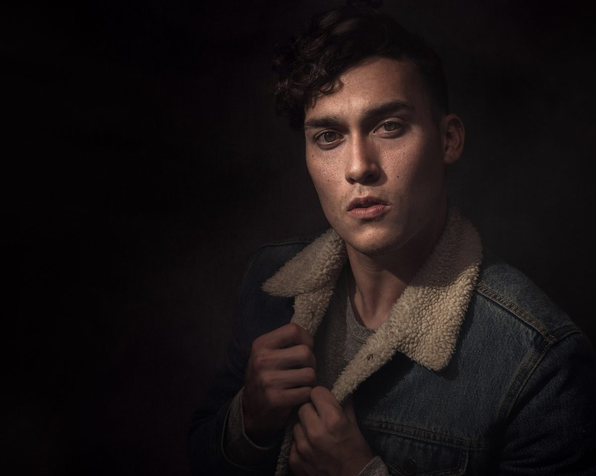 Young man in a shearling-collar denim jacket looking at the camera in moody studio lighting