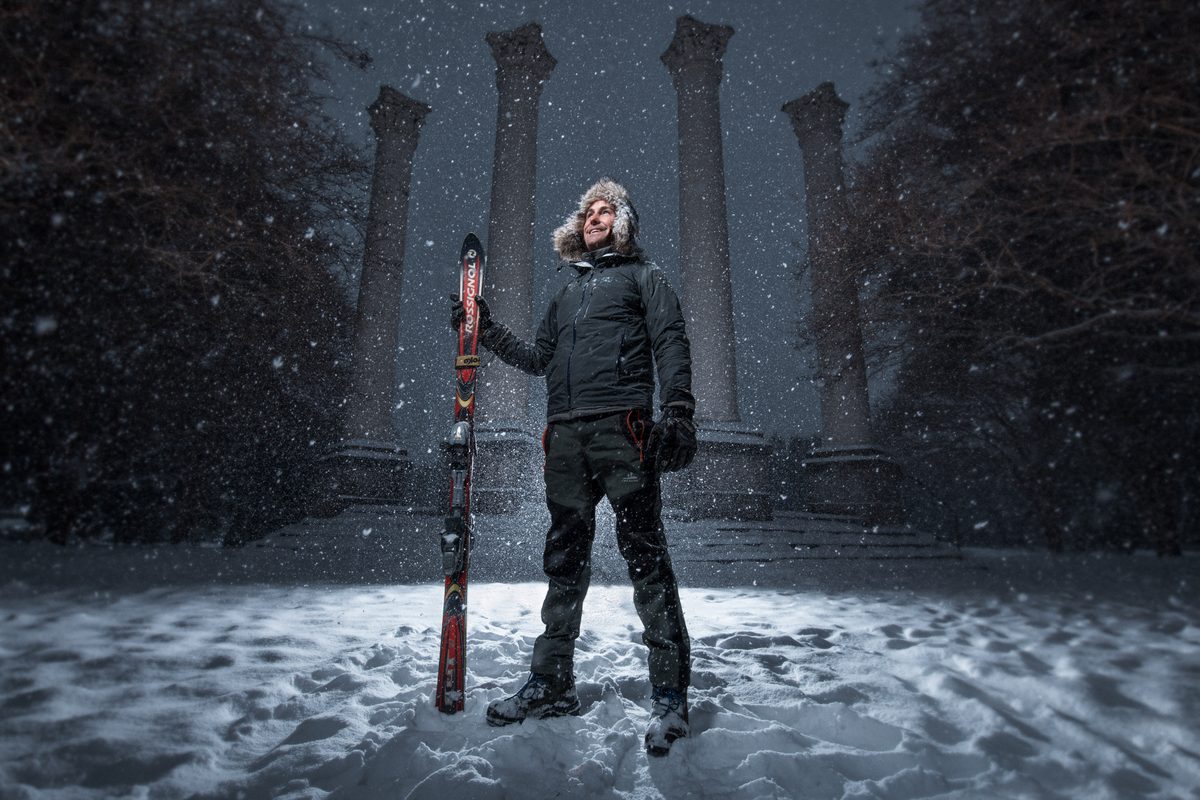 Man holding a pair of skis, standing in falling snow with a row of stone columns behind him