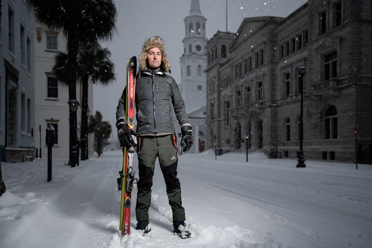 Man walking down a snow-covered street at night carrying skis, historic buildings and a church steeple behind him