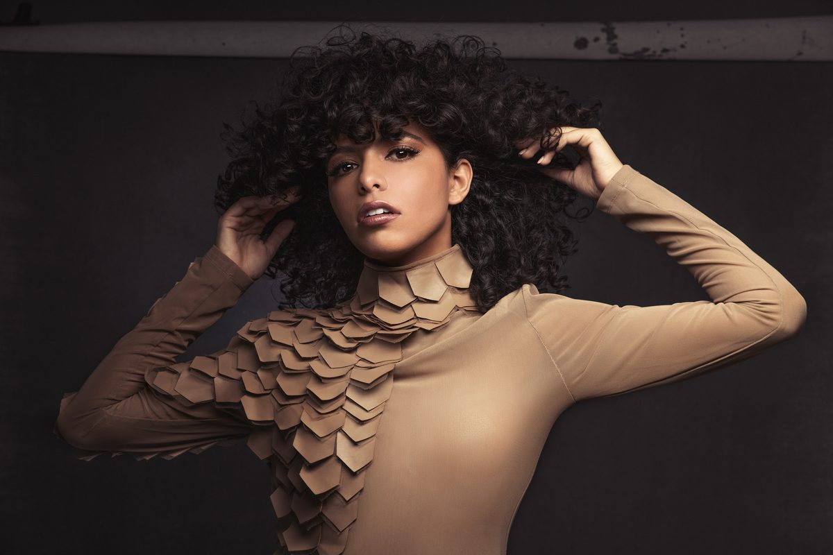 Woman with curly hair wearing a tan scale-textured top, hands in her hair, studio portrait
