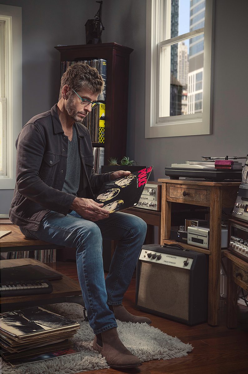 Man sitting on a stool playing a record in a room lined with vinyl records and stereo equipment