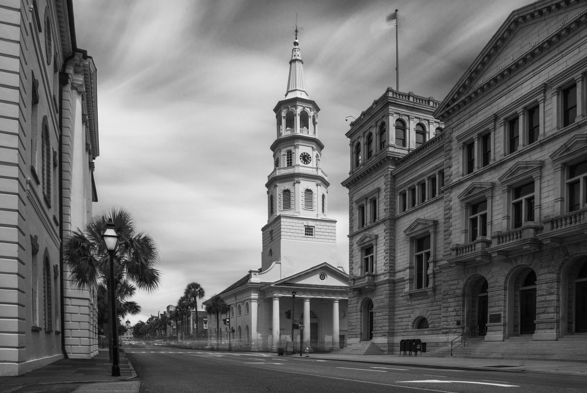 Black-and-white street view of a Charleston church steeple and colonial-era buildings along a palm-lined street