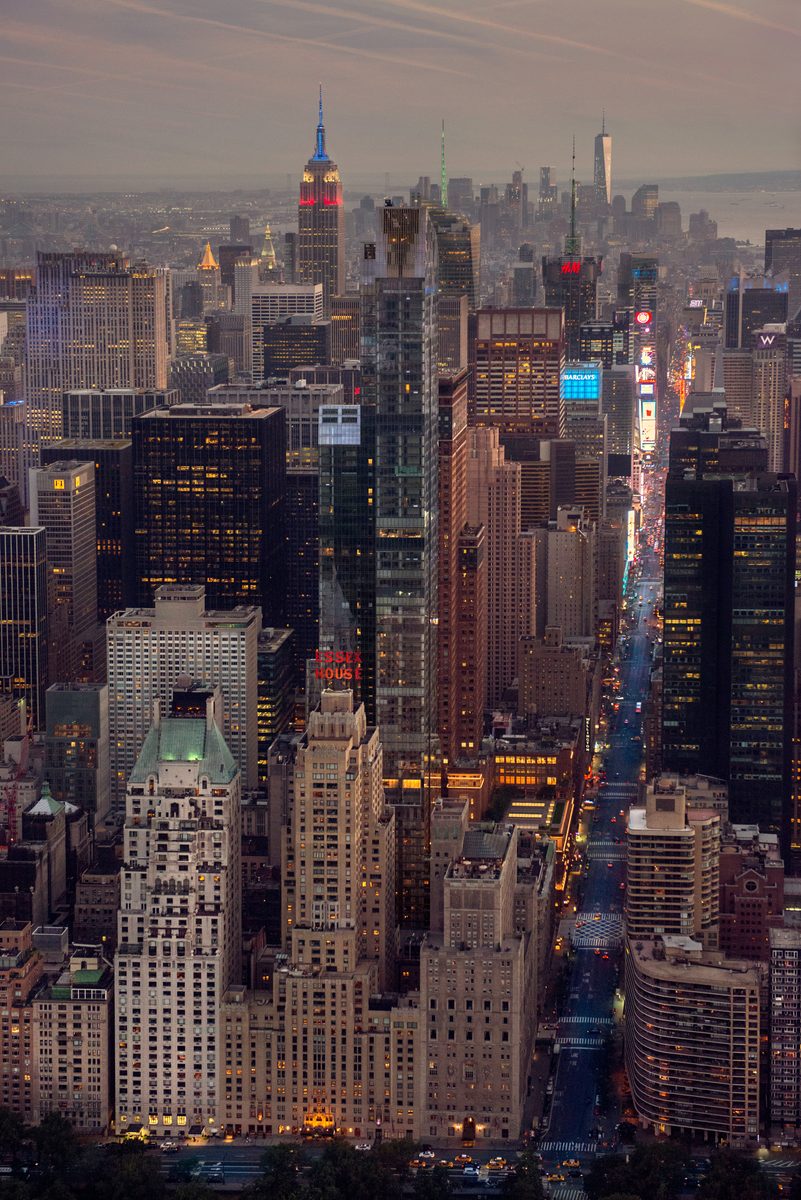 Aerial night view of Times Square and Midtown Manhattan skyscrapers lit up, with the Empire State Building visible in the distance
