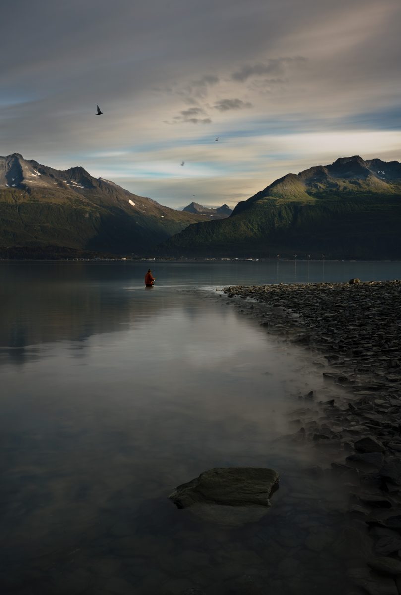 Calm lake at dusk with a person wading near shore and mountains reflected in the still water