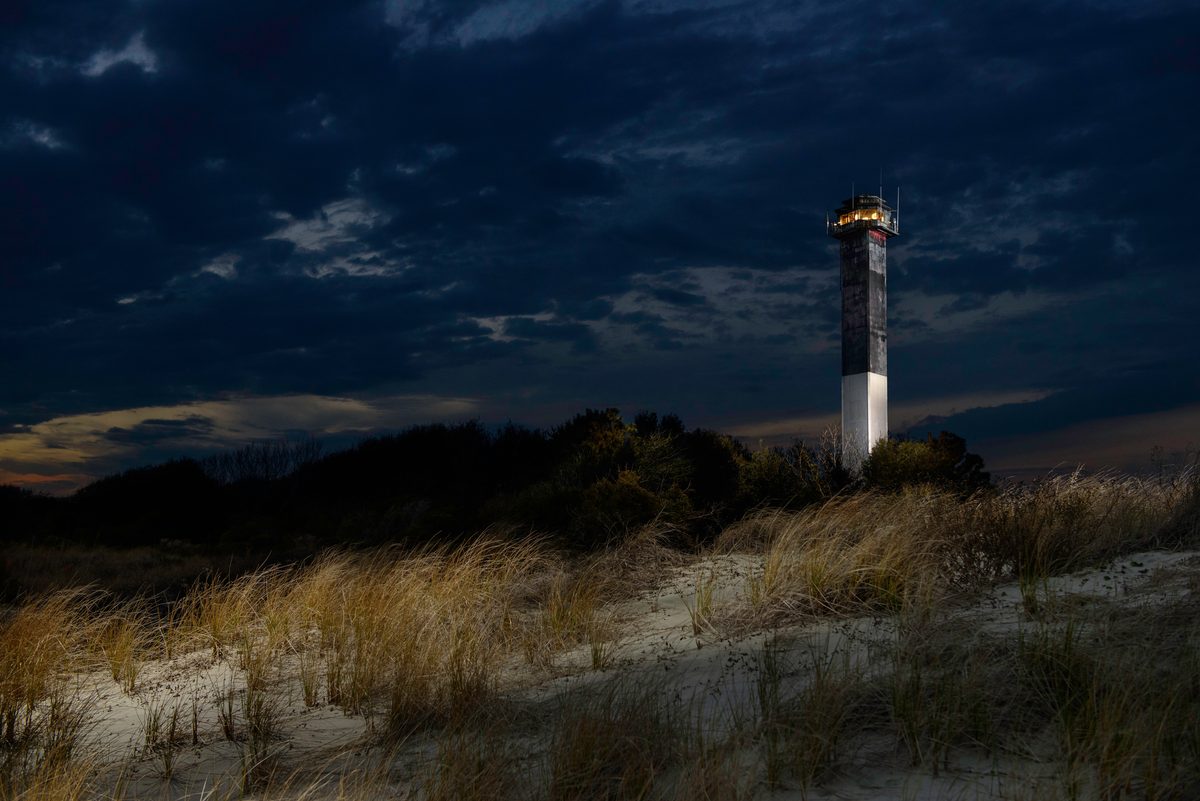 A lighthouse lit up at dusk beyond windswept dune grass under a darkening sky