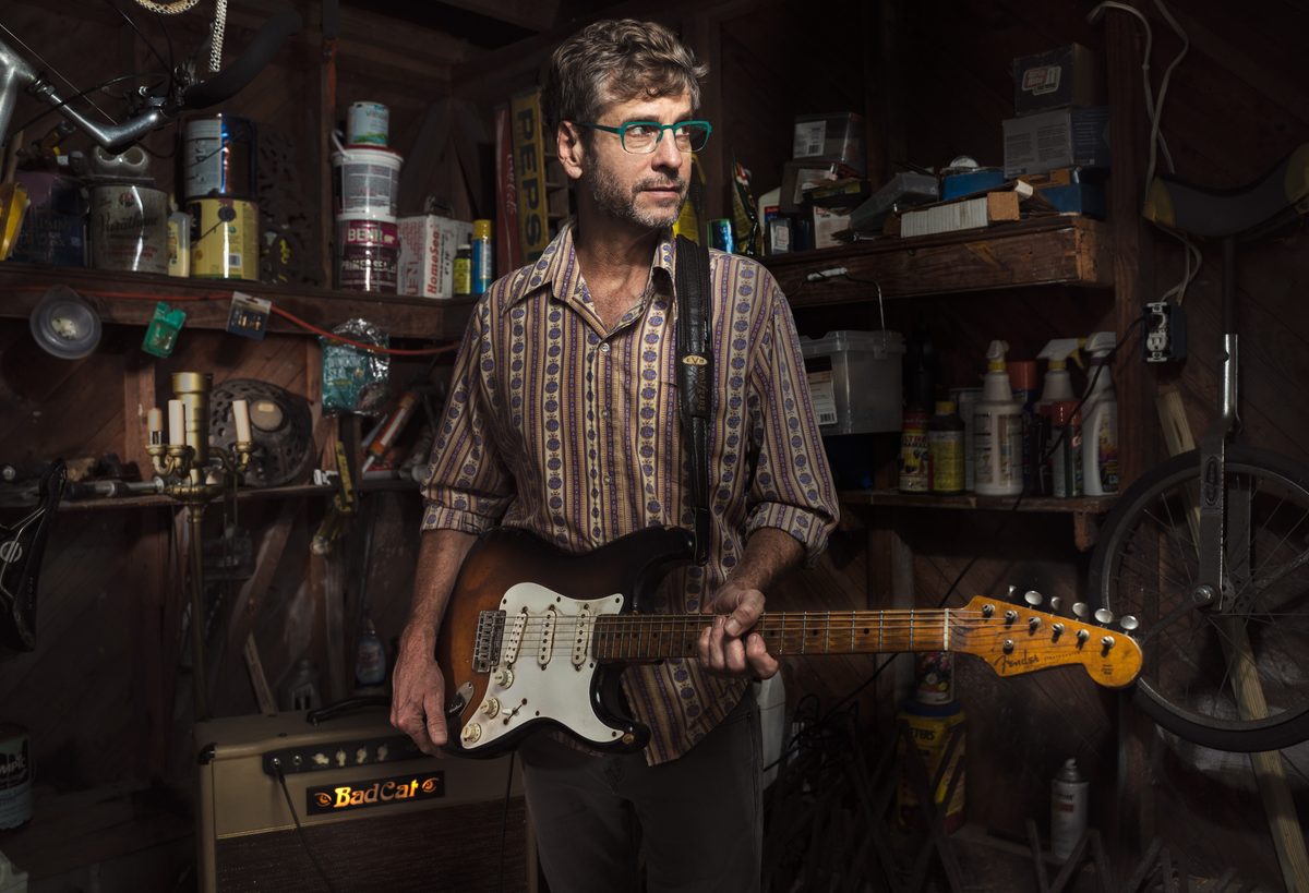 Man playing an electric guitar in a cluttered home workshop lined with tools and shelving