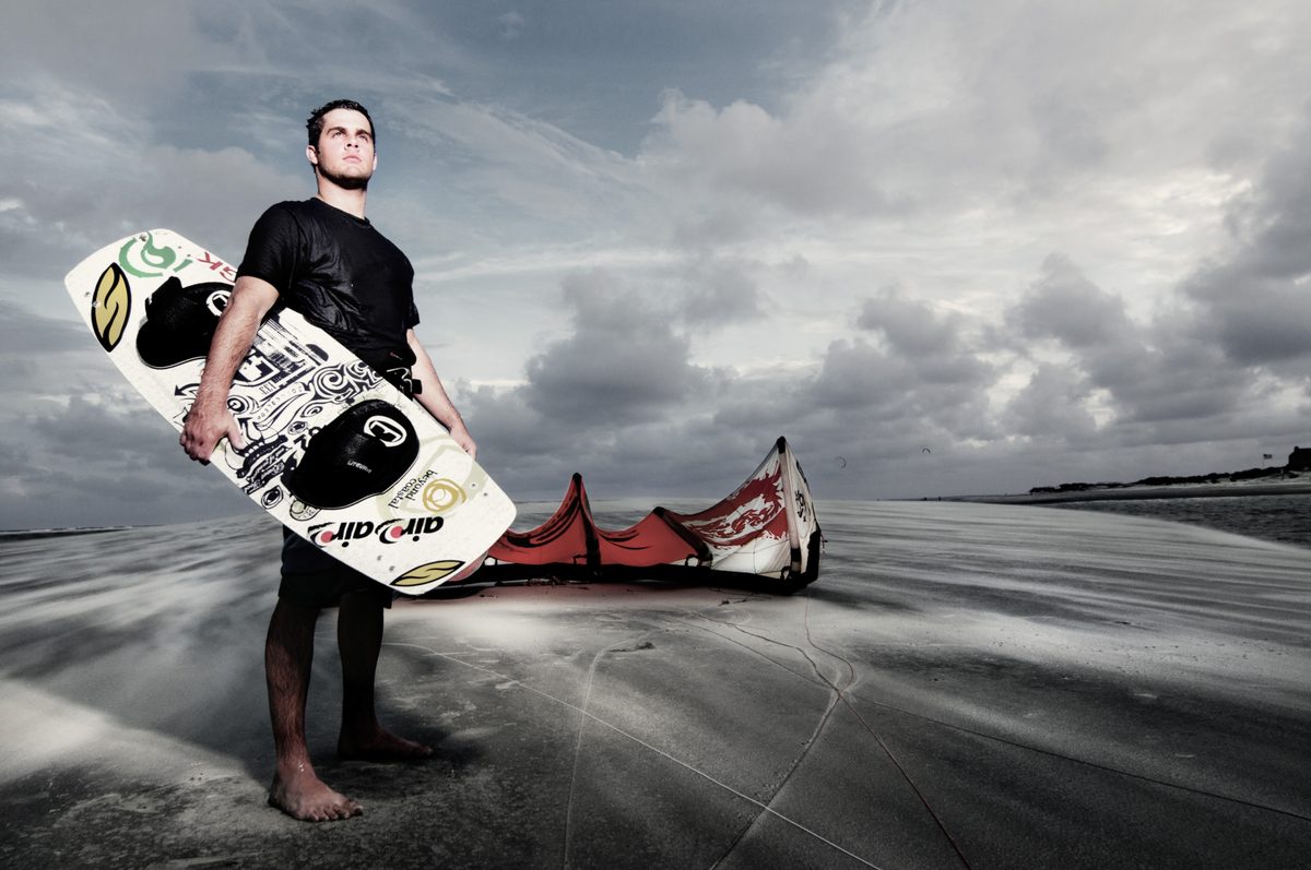 Man standing on a beach at dusk holding a wakeboard, kite laid out on the sand beside him