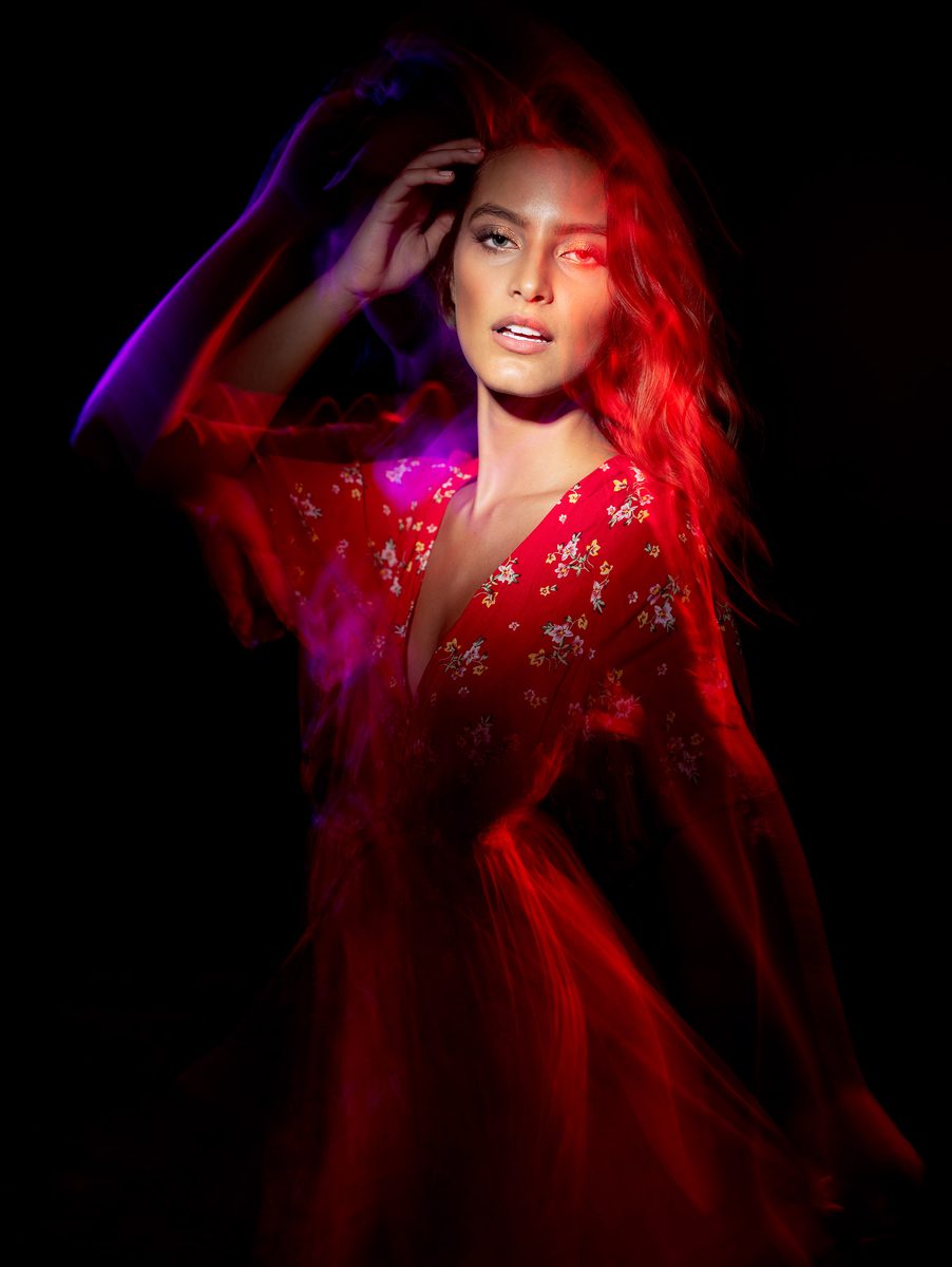 Woman in a red gown lit by dramatic colored light, hand in her hair