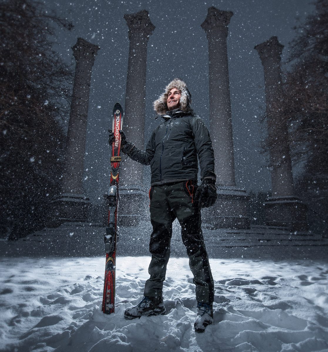 Man in winter gear holding a pair of skis, standing in falling snow in front of a columned building at night