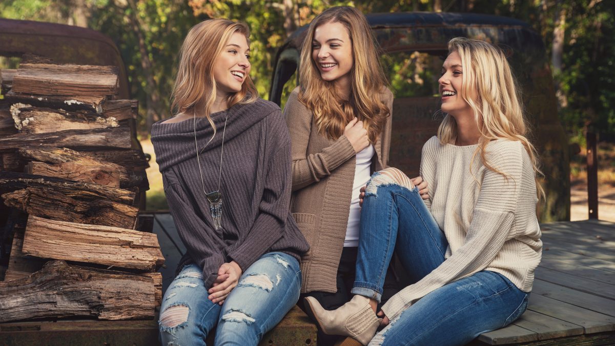 Three women sitting on the tailgate of an old pickup truck loaded with firewood, laughing together outdoors
