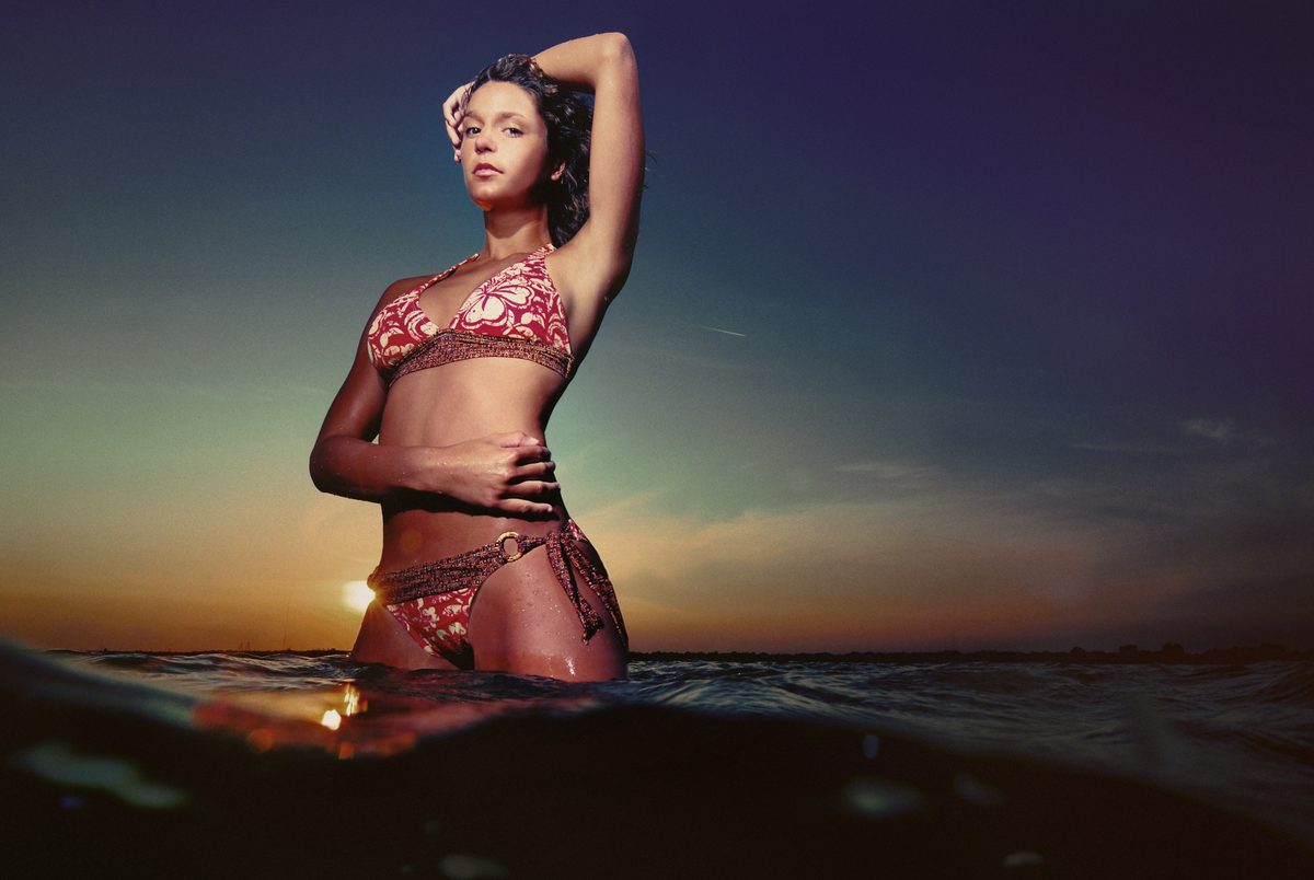Woman in a maroon bikini standing waist-deep in water at sunset, one hand in her hair