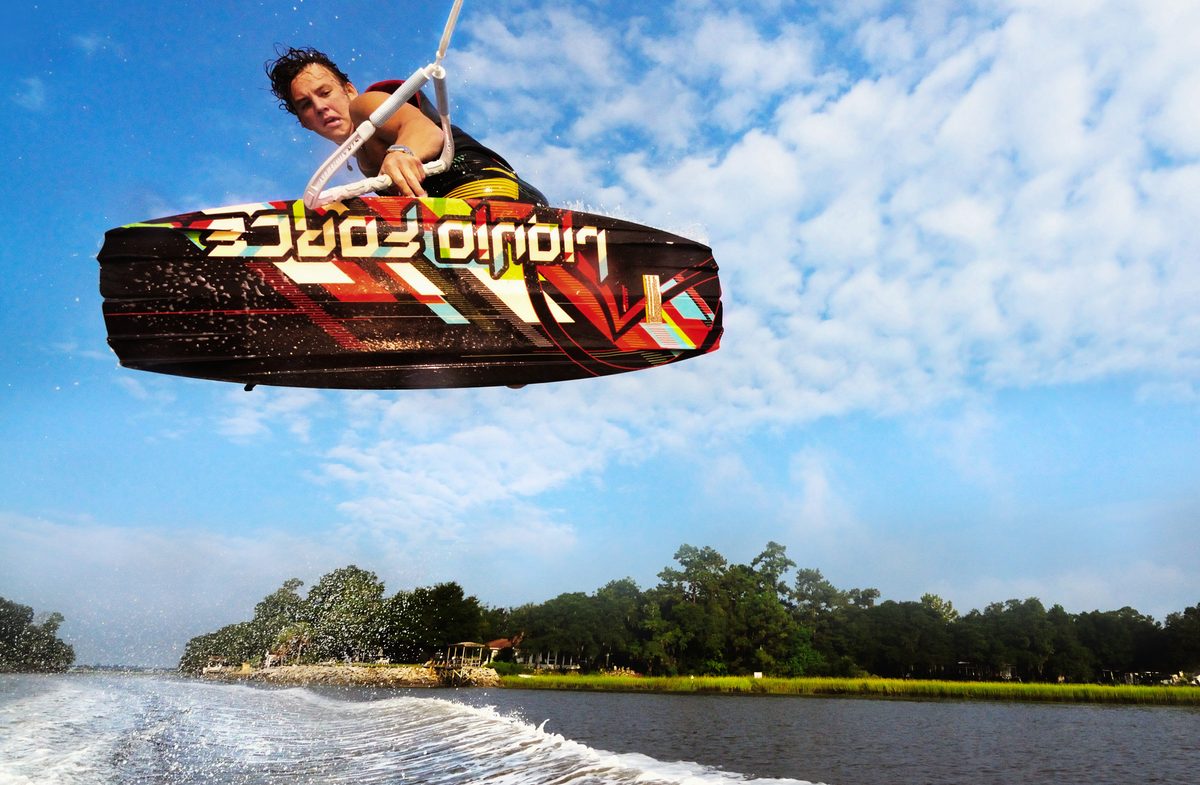 Wakeboarder flipping through a spray of water with trees lining the shore behind him