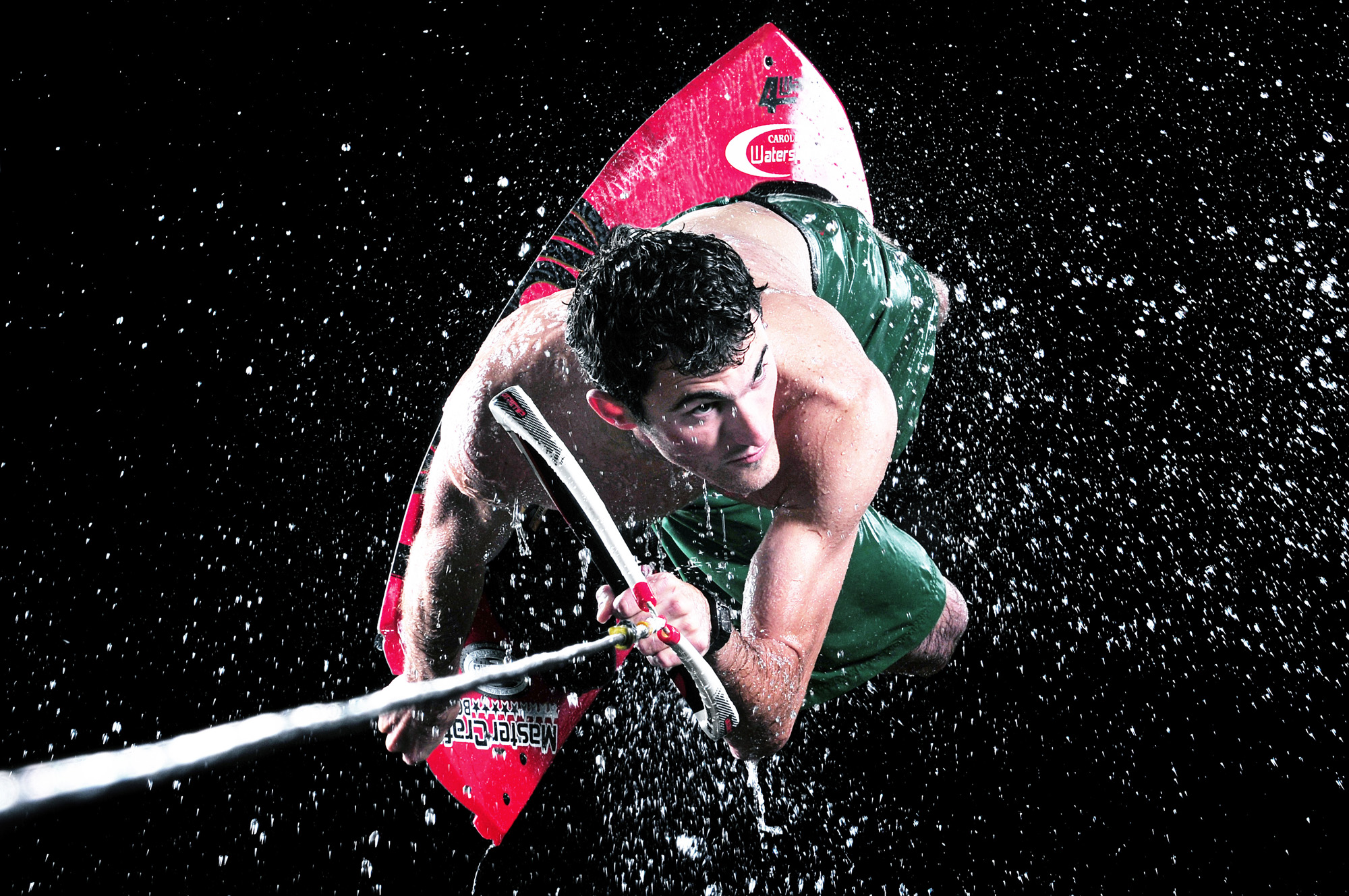 Man in a green top and red shorts gripping a boom, water splashing around him against a black background